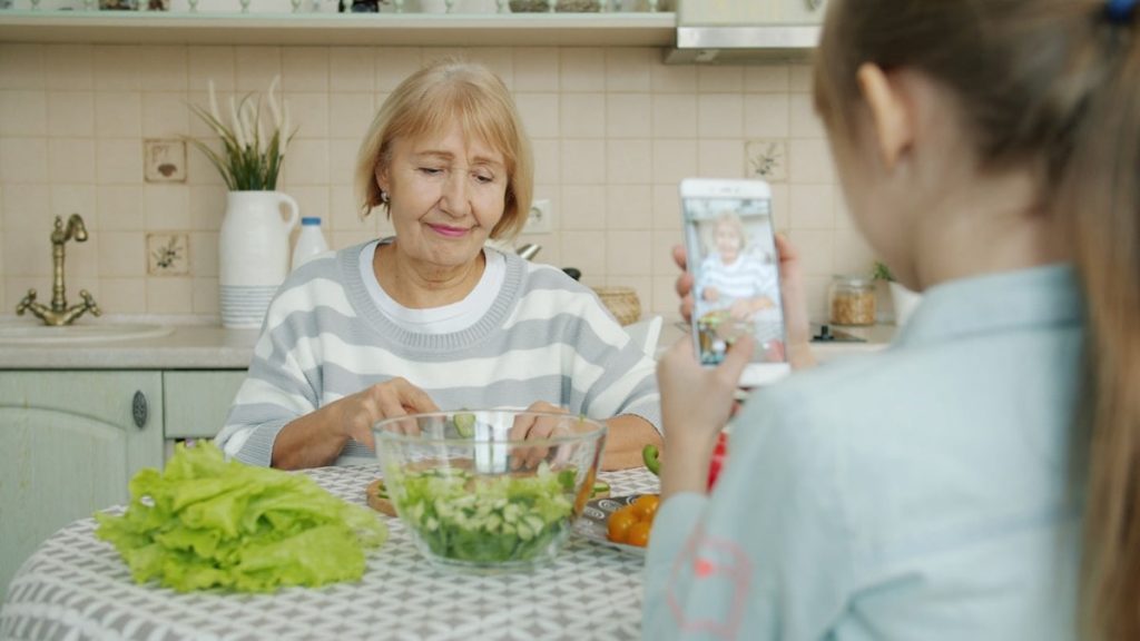 Little girl is taking pictures of granny who is cooking salad then posing for camera with cucumber slices having fun, child is using modern smartphone camera.